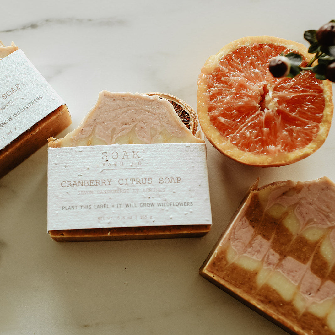 Three bars of soap with visible branding on a marble surface with dried oranges and flowers.