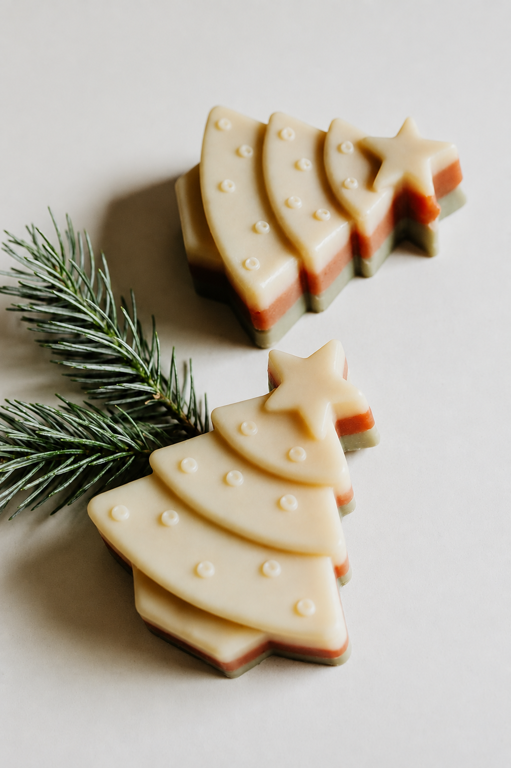 Three Christmas tree-shaped layered soaps in cream, orange, and green, styled with a pine branch on a light beige background.