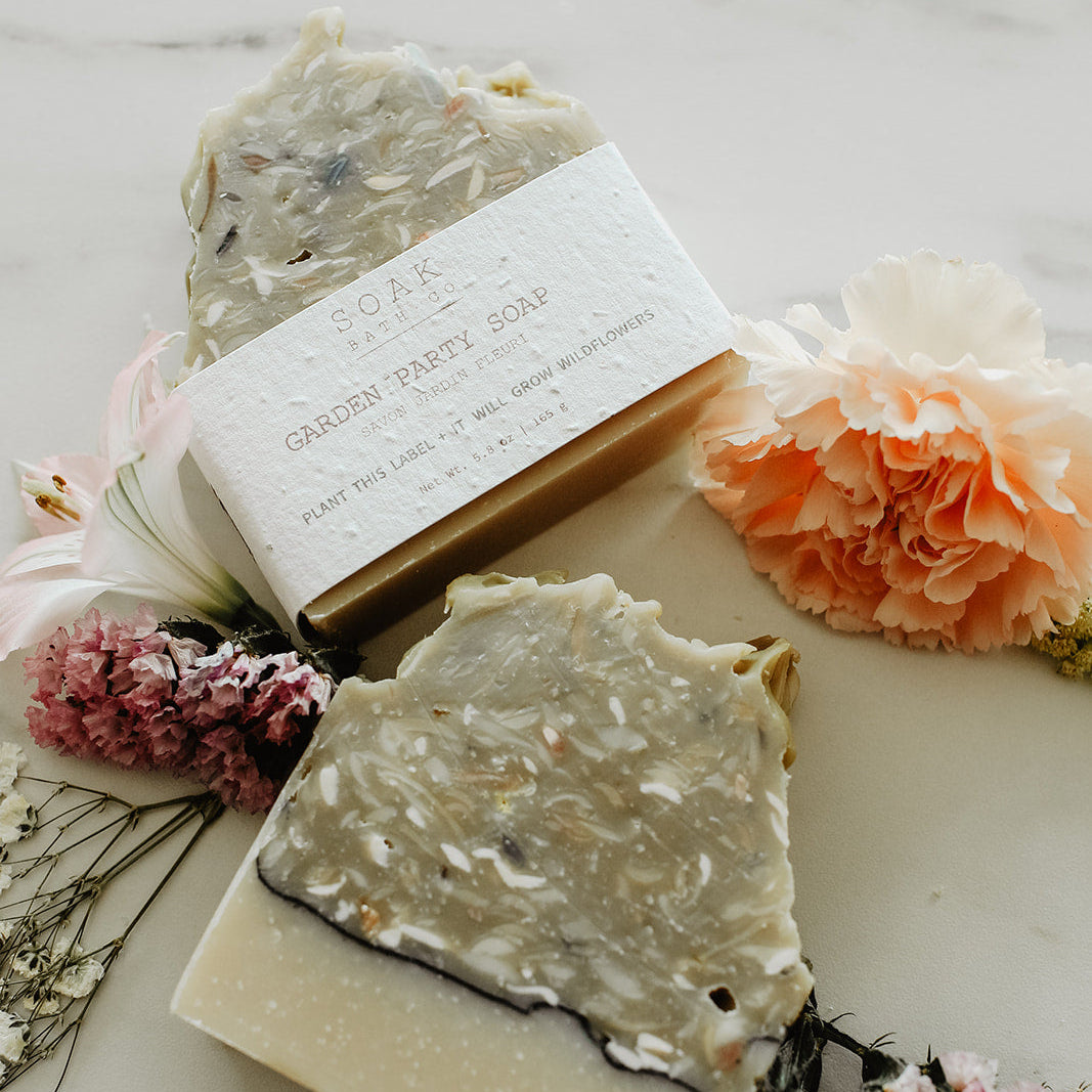 Two bars of soap with a box on a marble surface surrounded by flowers