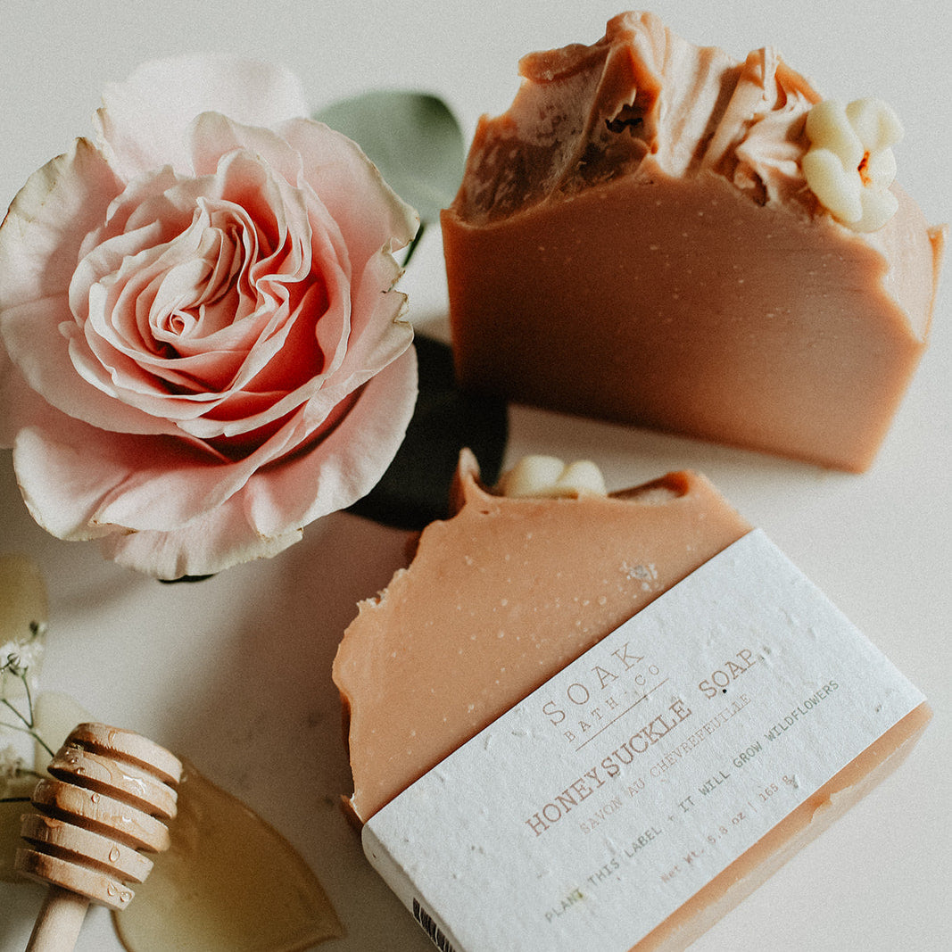Two bars of soap with a pink rose and honey dipper on a light surface