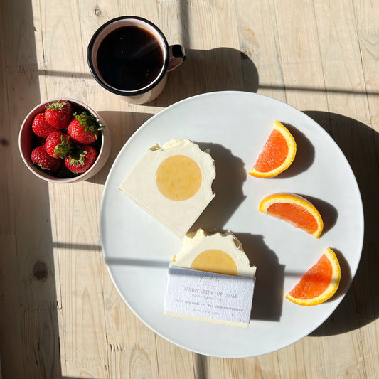 Plate with soap bar, orange wedges, and strawberries next to a cup of coffee on a wooden table.