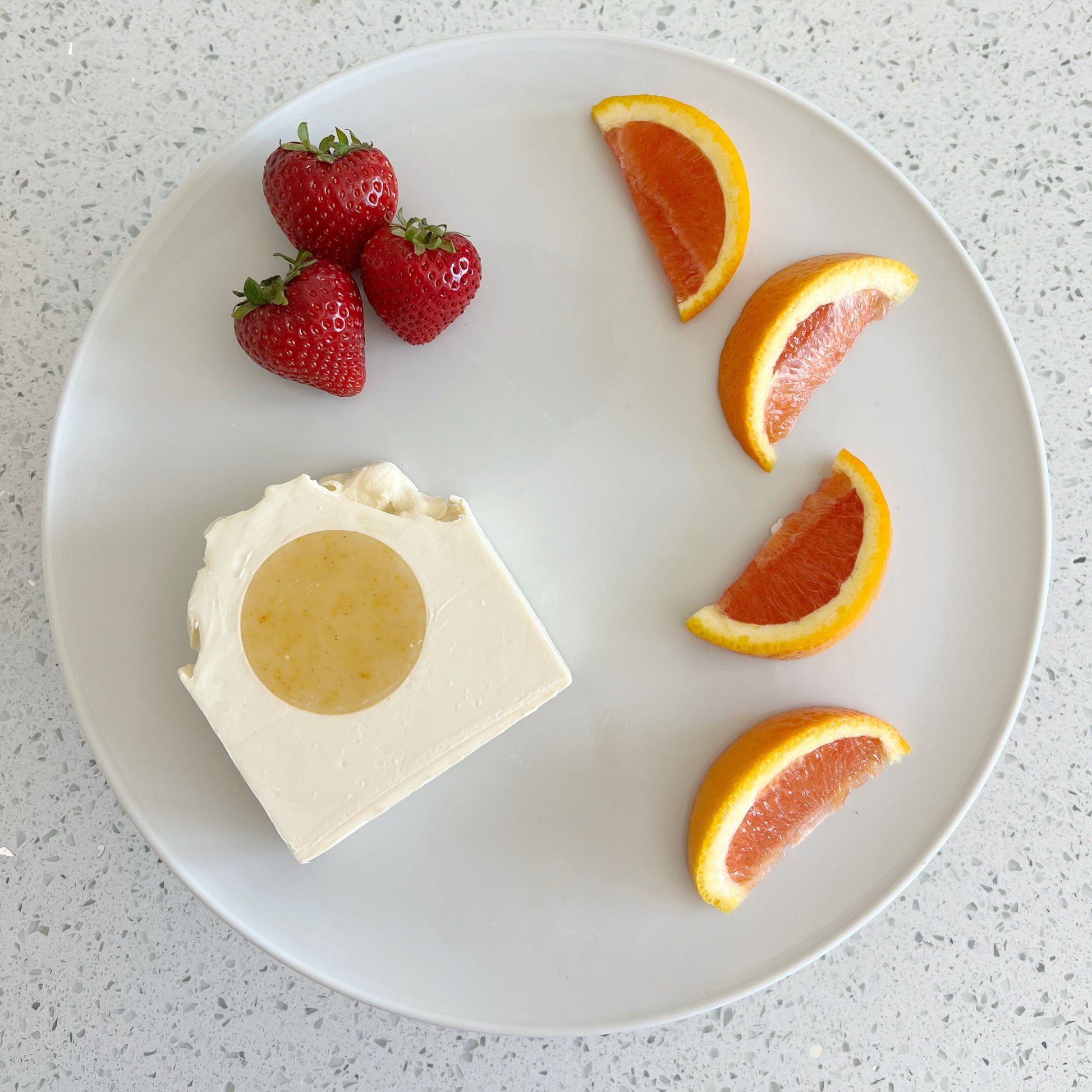 Soap bar with a round yellow center on a white plate with strawberries and grapefruit slices.