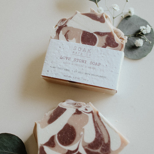 Two marbled soap bars with a 'Love Story Soap' packaging on a light background with green leaves.