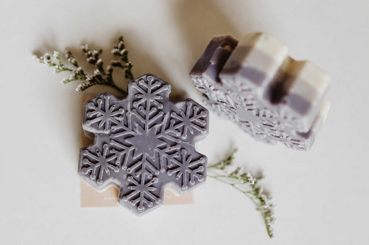 Two handmade soap bars with a snowflake design, one flat and one stacked, in a soft lavender-gray color, styled with small white flowers on a light beige background.