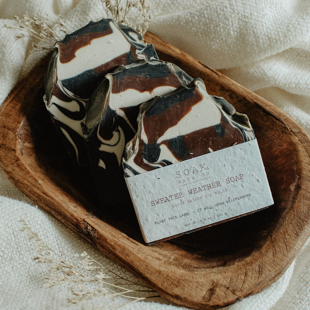 Three Sweater Weather soap bars with a decorative pattern in a wooden dish on a white fabric background