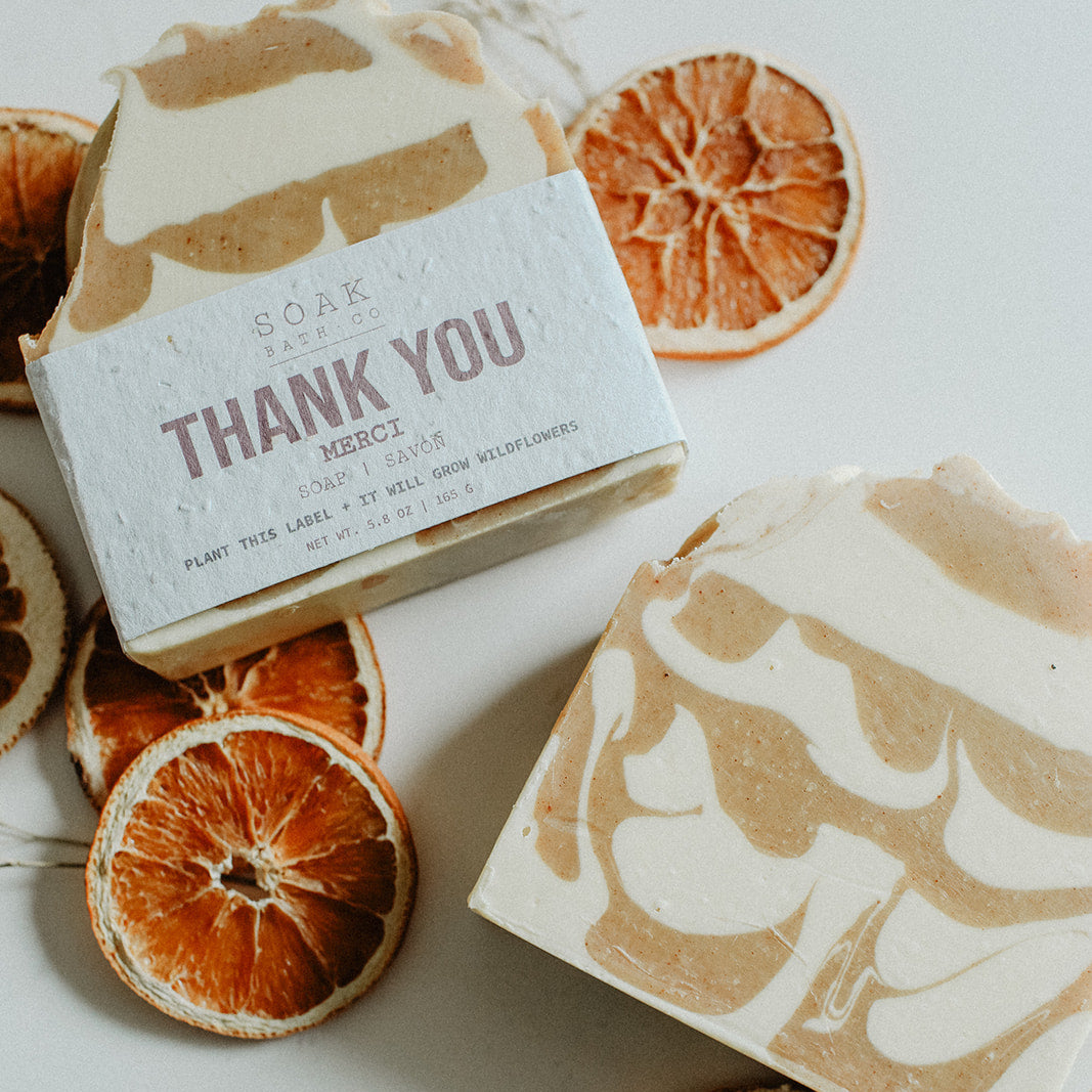 Two soap bars with decorative packaging on a white surface with dried orange slices.
