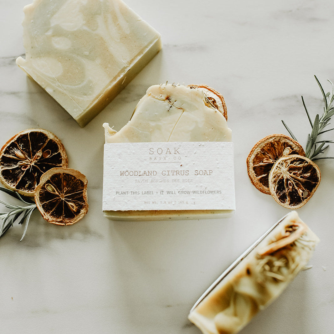 Three bars of soap with a label, surrounded by dried oranges and rosemary on a light surface.