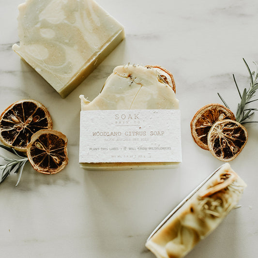 Three bars of soap with a label, surrounded by dried oranges and rosemary on a light surface.